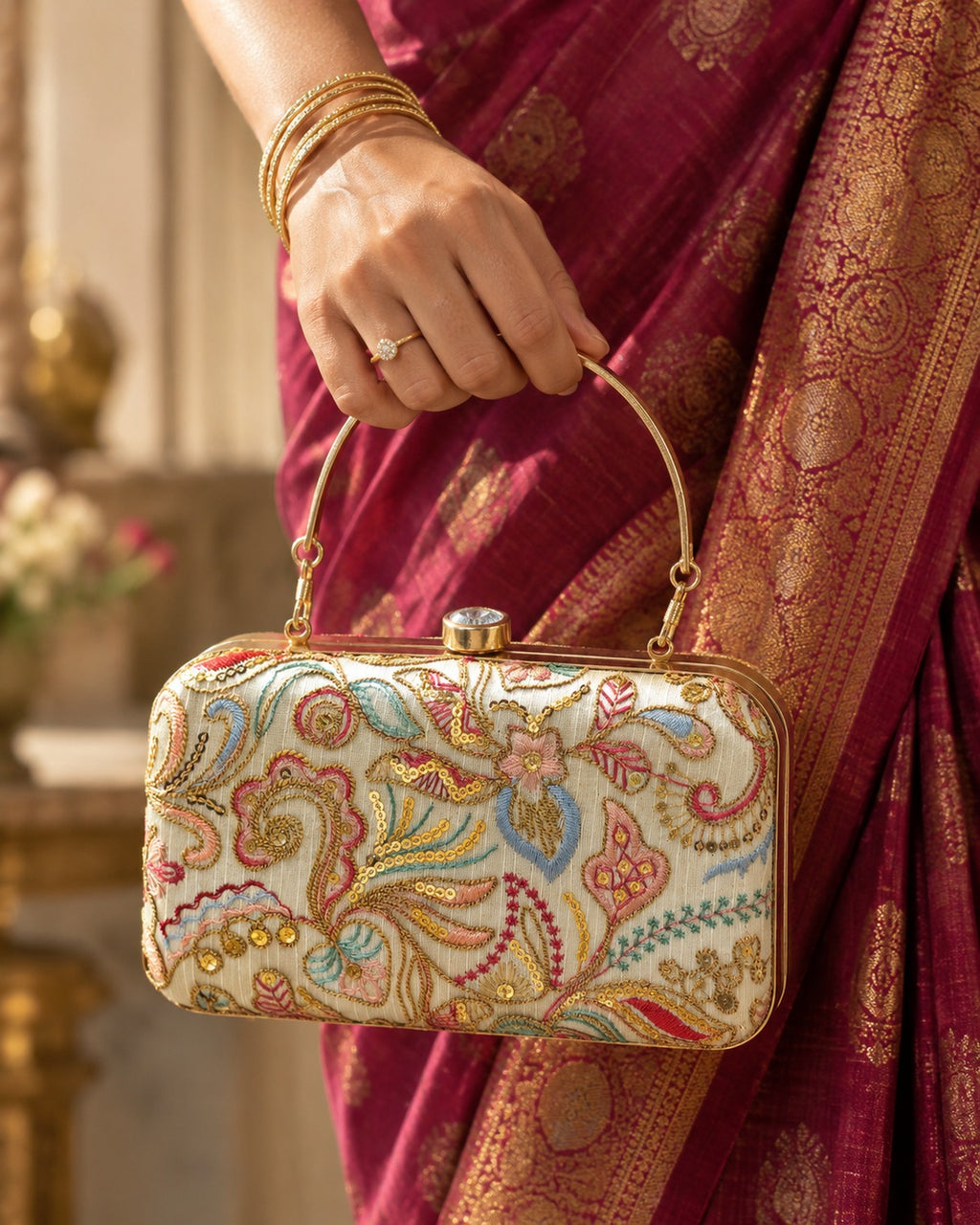 Embroidered box clutch with gold top handle, held against a silk saree